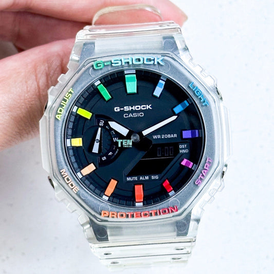 A hand-held CasiOak G-Shock watch with a colorful bezel featuring rainbow-like patterns and black dial with white casing and black hands.