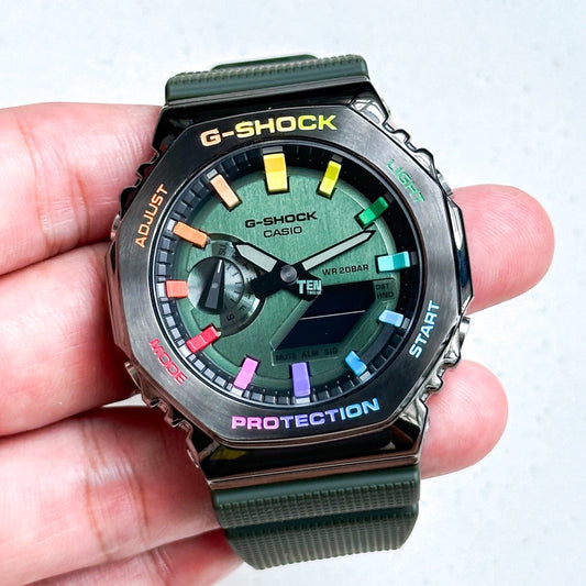 A hand holding a Casio G-Shock watch with a green band and a multicolored numeric bezel, displaying a digital screen with various colors and text such as 'G-SHOCK' and 'PROTECTION'.