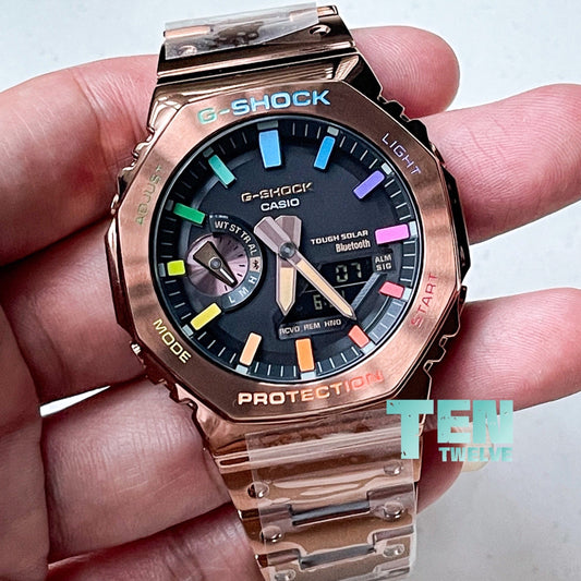 A person holding a rose gold CasiOak G-Shock watch with a multicolored rainbow pattern on the bezel and a clear crystal glass.