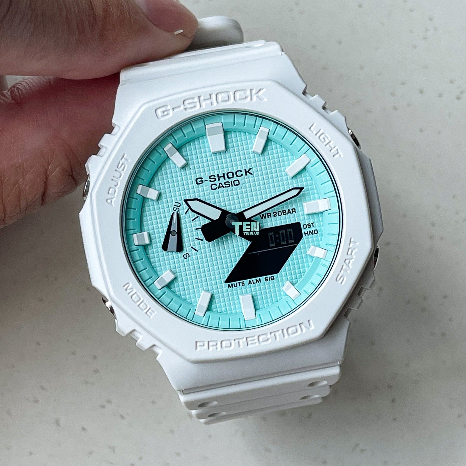 White Casio G-Shock watch with a turquoise dial held by a hand on a light gray background