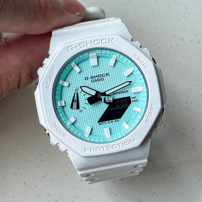 White Casio G-Shock watch with a turquoise dial held by a hand on a light gray background