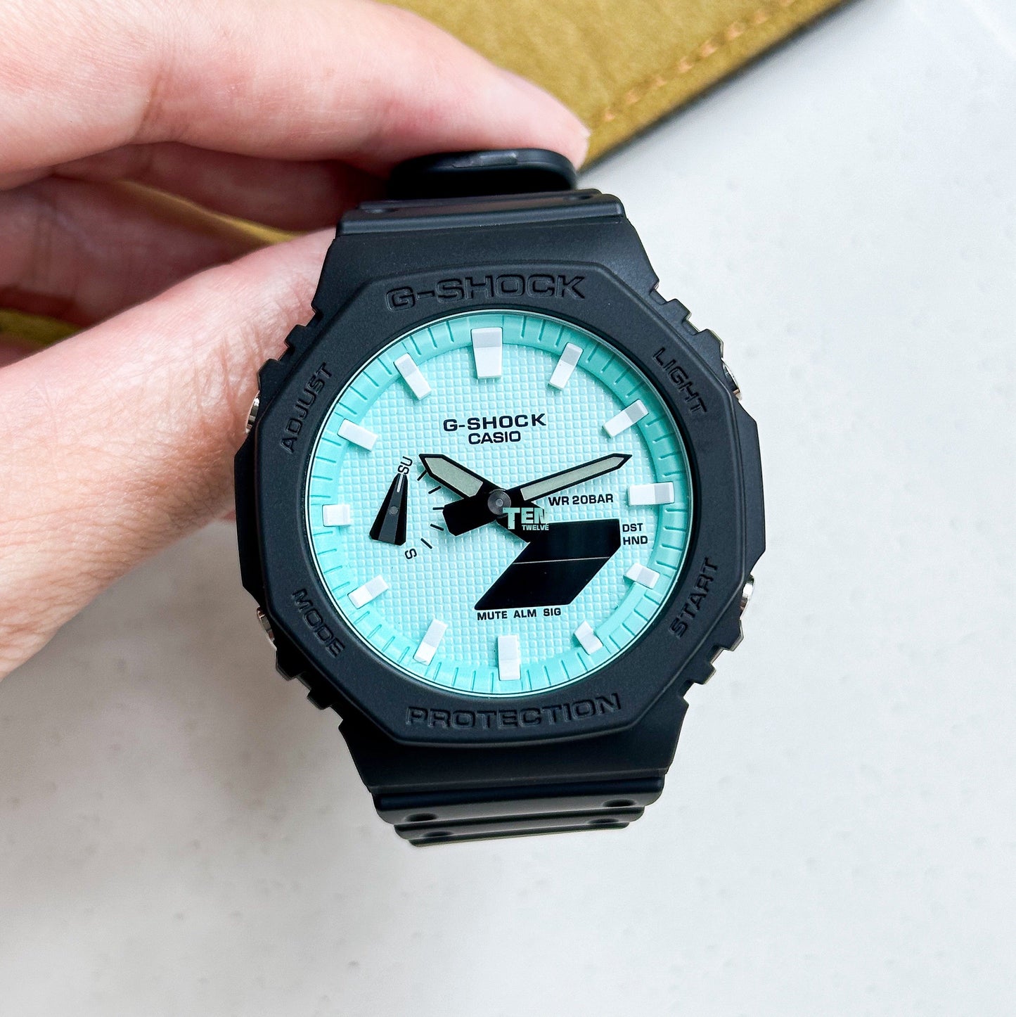 Black Casio G-Shock watch with a turquoise face held by a hand on a white background.
